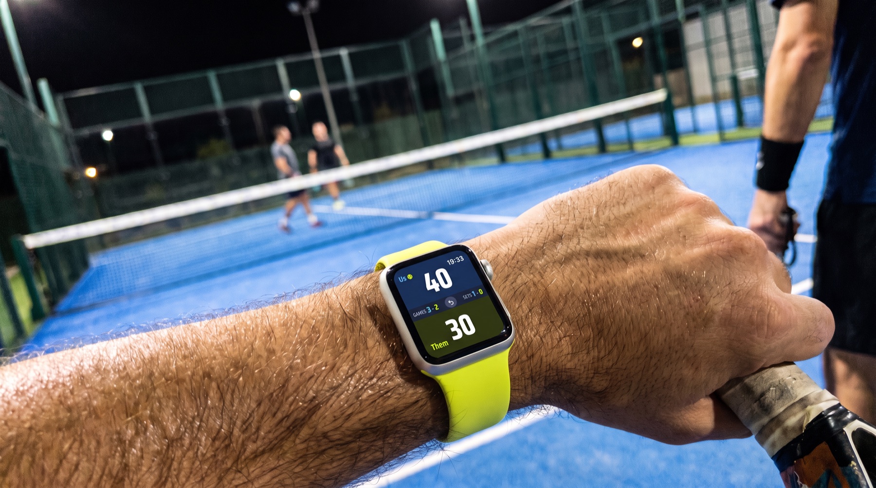 Poignet d'un joueur sur un court de padel le soir : l'Apple Watch affiche Padzo, Nous 40 au service, Eux 30, jeux 3-2, sets 1-0.